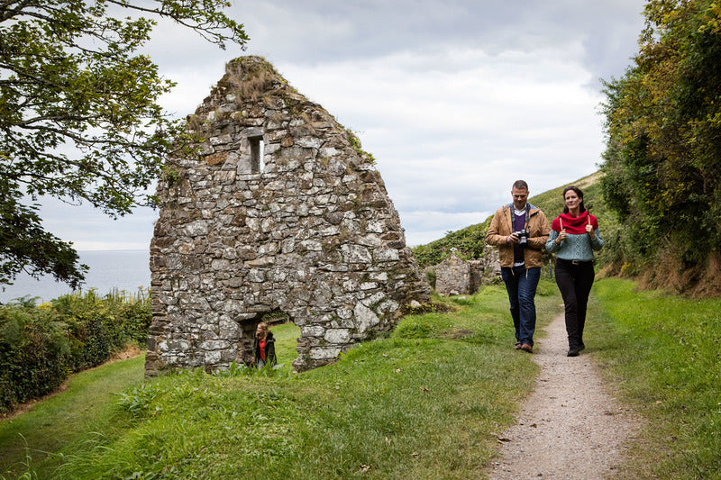 Two people walking on a path near an old stone building in a natural setting.